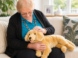 An elderly woman with short blonde hair and glasses sits on a beige sofa, gently petting a golden retriever weighted plush toy. She is wearing a turquoise blouse and a black cardigan.