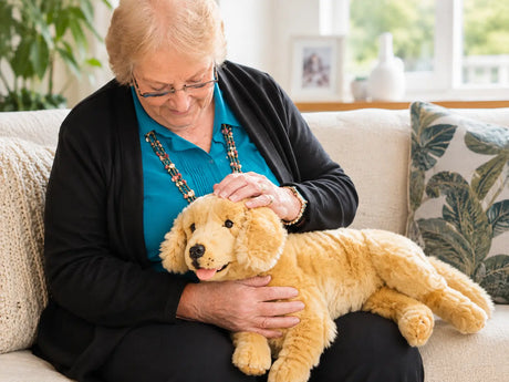 An elderly woman with short blonde hair and glasses sits on a beige sofa, gently petting a golden retriever weighted plush toy. She is wearing a turquoise blouse and a black cardigan.